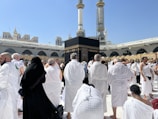 A large number of people, many dressed in white robes, are gathered around a black cubic structure with golden patterns and text. The background features a large open courtyard with arches and tall minarets under a clear blue sky. Some individuals appear to be praying, while others are walking or standing.