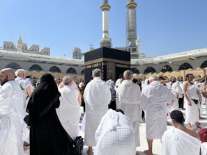 A large number of people, many dressed in white robes, are gathered around a black cubic structure with golden patterns and text. The background features a large open courtyard with arches and tall minarets under a clear blue sky. Some individuals appear to be praying, while others are walking or standing.