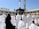 A large number of people, many dressed in white robes, are gathered around a black cubic structure with golden patterns and text. The background features a large open courtyard with arches and tall minarets under a clear blue sky. Some individuals appear to be praying, while others are walking or standing.