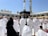 A large number of people, many dressed in white robes, are gathered around a black cubic structure with golden patterns and text. The background features a large open courtyard with arches and tall minarets under a clear blue sky. Some individuals appear to be praying, while others are walking or standing.
