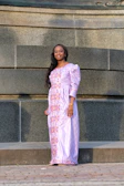 A stylish woman in a flowing lavender dress posing against a cream-colored wall with soft pink floral accents.
