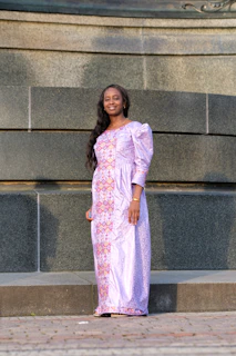 A young woman in a soft lavender dress smiling confidently against a pastel pink background.