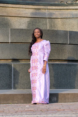 A stylish woman in a flowing lavender dress posing against a cream-colored wall with soft pink floral accents.