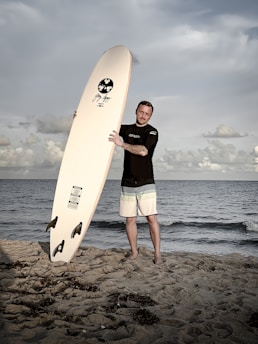 A person holding a large surfboard stands on a sandy beach with the ocean in the background. The sky is partly cloudy and the waves are gentle. The person is wearing a wetsuit and shorts.