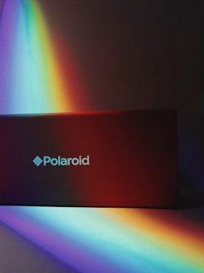 A rectangular object with the 'Polaroid' logo is bathed in a vibrant spectrum of rainbow colors. The interplay of light creates a vivid and colorful display across the surface, blending gradually from deep blues and purples to warm oranges and reds.