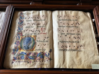 An illuminated manuscript featuring ornate calligraphy and neumes on a two-page spread. The left page is adorned with decorative floral motifs and an elaborate initial containing a painted figure. The right page contains text with musical notation on red lines.