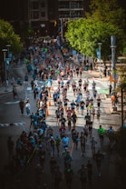 A large group of people are participating in a marathon, running down a city street surrounded by spectators and trees. The street is marked with cones and there are various buildings in the background.