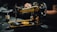A vintage sewing machine with ornate gold and black detailing is positioned on a dark surface. The intricate patterns and the name 'Sinclair' are visible on the machine. In the background, there is a blurred figure, suggesting a person operating or examining the machine. The setting appears dimly lit, focusing on the sewing machine's craftsmanship.