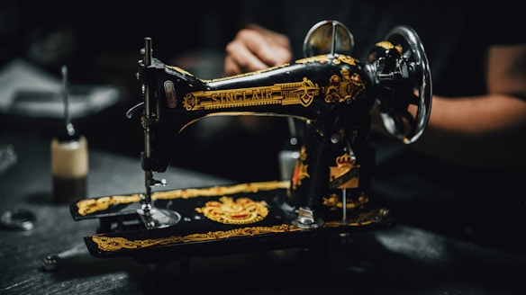 A vintage sewing machine with ornate gold and black detailing is positioned on a dark surface. The intricate patterns and the name 'Sinclair' are visible on the machine. In the background, there is a blurred figure, suggesting a person operating or examining the machine. The setting appears dimly lit, focusing on the sewing machine's craftsmanship.