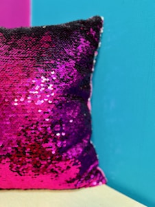 A close-up of a sequined decorative pillow with sparkling pink and purple sequins. The pillow is set against a bright turquoise wall with a small portion of a wooden surface visible at the bottom.