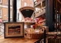 A rustic indoor scene features an old-fashioned weighing scale surrounded by various packaged goods, including bags of snacks and nuts. A framed picture rests on a wooden table, next to a wicker basket filled with baked goods. The setup is against a large window overlooking a street with brick buildings and a parked red car.