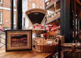 A rustic indoor scene features an old-fashioned weighing scale surrounded by various packaged goods, including bags of snacks and nuts. A framed picture rests on a wooden table, next to a wicker basket filled with baked goods. The setup is against a large window overlooking a street with brick buildings and a parked red car.