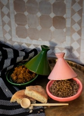 A gold-toned tray is set against a geometrically patterned tile wall. On the tray, two tagine pots are present, one green and one pink, containing a lentil dish and a seasoned dish respectively. A piece of flatbread is placed alongside a wooden spoon, all resting on a black fabric with white patterns.