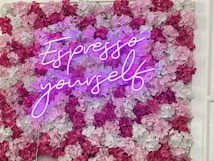 A neon sign with the phrase 'Espresso Yourself' is mounted on a vibrant floral wall composed of pink, red, and white flowers. The neon sign's purple glow creates a striking contrast against the textured background.