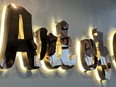 Glossy, reflective 3D black letters mounted on a wall, illuminated from behind with a warm light, casting soft shadows. The surface of the letters reflects surrounding colors and shapes, creating a dynamic visual effect.