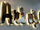 Glossy, reflective 3D black letters mounted on a wall, illuminated from behind with a warm light, casting soft shadows. The surface of the letters reflects surrounding colors and shapes, creating a dynamic visual effect.