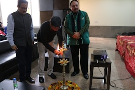 Three men are attending a ceremonial event indoors. One man is lighting a brass lamp placed on a decorated stand. The setting includes chairs, a red draped table, and a projector on a small table.