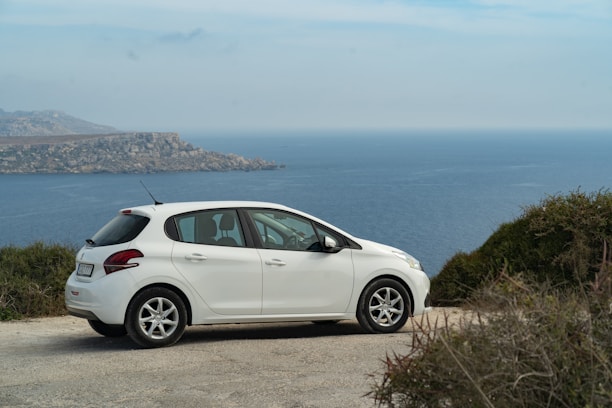 A white hatchback car is parked on a gravel road overlooking a vast ocean with a clear horizon and distant rocky cliffs. Surrounding the viewpoint are patches of green shrubbery.