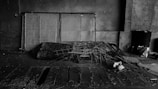 A dimly lit, industrial setting featuring a collection of metal grids leaning against a concrete wall. There are scattered pieces of debris and a pile of intertwined wires and scrap metal on the floor. The area has an abandoned and neglected feel, with some papers and a black barrel visible in the background.