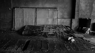 A dimly lit, industrial setting featuring a collection of metal grids leaning against a concrete wall. There are scattered pieces of debris and a pile of intertwined wires and scrap metal on the floor. The area has an abandoned and neglected feel, with some papers and a black barrel visible in the background.