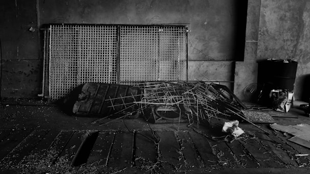 A dimly lit, industrial setting featuring a collection of metal grids leaning against a concrete wall. There are scattered pieces of debris and a pile of intertwined wires and scrap metal on the floor. The area has an abandoned and neglected feel, with some papers and a black barrel visible in the background.