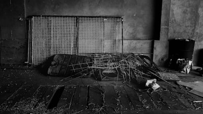A dimly lit, industrial setting featuring a collection of metal grids leaning against a concrete wall. There are scattered pieces of debris and a pile of intertwined wires and scrap metal on the floor. The area has an abandoned and neglected feel, with some papers and a black barrel visible in the background.
