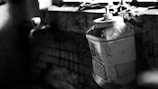 A large plastic container with a pump sits on a dirty, cracked windowsill with grime and cobwebs. The surrounding environment appears neglected, with visible decay and darkness dominating the scene.