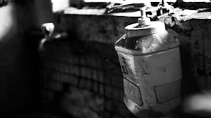 A large plastic container with a pump sits on a dirty, cracked windowsill with grime and cobwebs. The surrounding environment appears neglected, with visible decay and darkness dominating the scene.