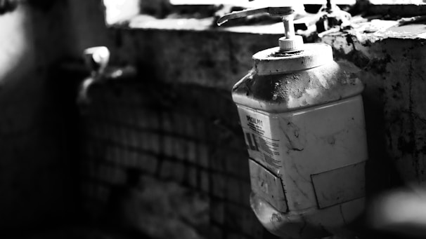 A large plastic container with a pump sits on a dirty, cracked windowsill with grime and cobwebs. The surrounding environment appears neglected, with visible decay and darkness dominating the scene.
