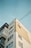 The image depicts a multi-story residential building with a minimalist architectural style. The structure features a combination of white and beige sections, with several windows. Visible power lines intersect the scene above the building, creating a network against a clear, pale blue sky.