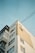 The image depicts a multi-story residential building with a minimalist architectural style. The structure features a combination of white and beige sections, with several windows. Visible power lines intersect the scene above the building, creating a network against a clear, pale blue sky.