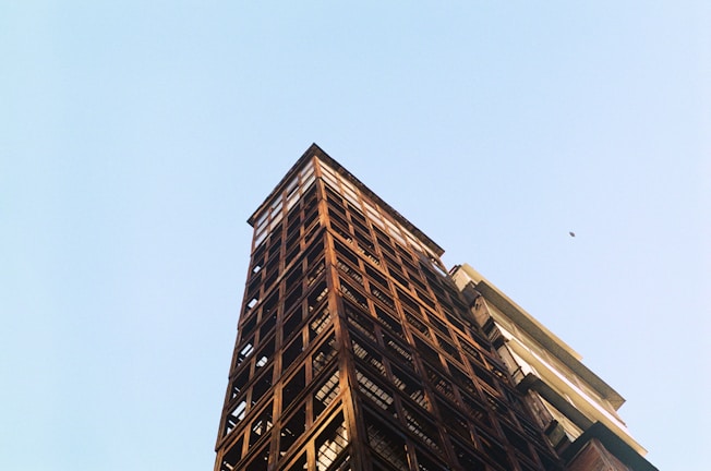A tall, unfinished building structure with a steel frame and an open-air design extends upwards into a clear blue sky. The perspective emphasizes the height and angular geometry of the architecture, suggesting a sense of rise and aspiration.