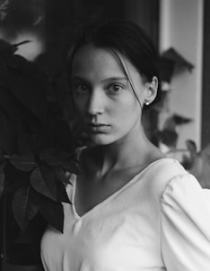 A young woman with a neutral expression is gazing towards the camera. She is wearing a light-colored top and has her hair pulled back. Lush foliage is visible in the background, contributing to a classic and serene composition.