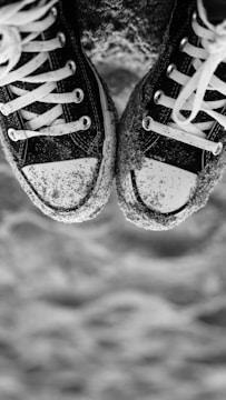 A pair of black and white sneakers with white laces covered in sand. The sneakers are positioned close together, and the background appears to be a sandy surface, possibly a beach.