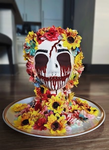 A decorative skull is covered with a variety of colorful flowers, including sunflowers and roses, and is artistically painted with black and red drips resembling tears. It sits on a dish with additional flowers surrounding it, creating a vibrant display.