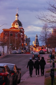 A streetscape features a prominent historic building with a dome and clock tower, surrounded by other architectural structures. The street is lined with parked cars and trees, and a group of three people walks down the sidewalk. The scene is set during golden hour, casting a warm glow on the buildings.