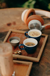 A wooden tray on a rustic table holds three cups containing different beverages, including an espresso in a blue cup. A small bowl of milk and a golden spoon accompany the cups. Large, over-ear headphones rest nearby, suggesting a cozy and relaxing atmosphere.