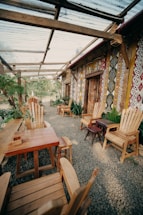 A rustic outdoor seating area with wooden furniture arranged on a gravel floor. The space features multiple chairs and tables made of natural wood, surrounded by lush greenery. The wall is decorated with colorful, patterned artwork. A transparent roof made of panels provides partial shade, supported by wooden beams.