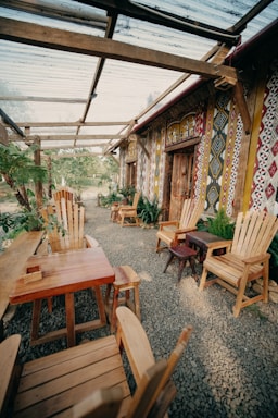 A rustic outdoor seating area with wooden furniture arranged on a gravel floor. The space features multiple chairs and tables made of natural wood, surrounded by lush greenery. The wall is decorated with colorful, patterned artwork. A transparent roof made of panels provides partial shade, supported by wooden beams.