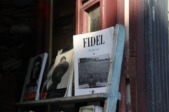A stack of magazines on a wooden shelf with a focus on a magazine featuring the title 'FIDEL' in bold letters. The cover shows a black and white photograph of a large crowd with a prominent figure looking over them from the forefront. The shelf is positioned next to a window, and the lighting creates shadows across the cover.
