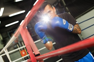 A man is standing in a boxing ring wearing blue boxing gloves. He appears to be training or preparing for a match. The setting seems to be a well-equipped gym with visible boxing ropes and a blurred background.