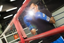 A man is standing in a boxing ring wearing blue boxing gloves. He appears to be training or preparing for a match. The setting seems to be a well-equipped gym with visible boxing ropes and a blurred background.