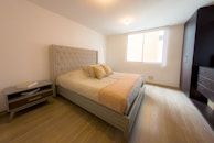 A modern bedroom with a large bed featuring a tufted headboard and several pillows. The bed is dressed with a light-colored quilt. There is a nightstand on one side with books placed on top. The room has wooden flooring and a large window allowing natural light to illuminate the space. A dark-colored wardrobe is positioned on one side of the room.