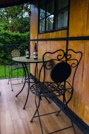 A cozy outdoor patio with a wooden wall, featuring a small round mosaic table and two decorative wrought iron chairs. Two glass bottles are placed on the table, and the area is bordered by lush green bushes.