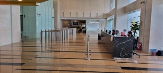 A spacious lobby area with polished floors and a row of modern turnstiles leading to a corridor. On the right, a long black marble reception desk with several people sitting and working. Glass walls and indoor plants enhance the open and bright atmosphere.