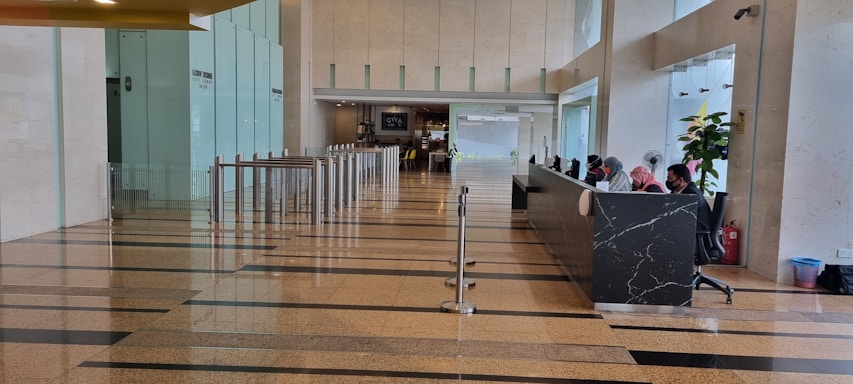 A spacious lobby area with polished floors and a row of modern turnstiles leading to a corridor. On the right, a long black marble reception desk with several people sitting and working. Glass walls and indoor plants enhance the open and bright atmosphere.