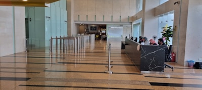 A spacious lobby area with polished floors and a row of modern turnstiles leading to a corridor. On the right, a long black marble reception desk with several people sitting and working. Glass walls and indoor plants enhance the open and bright atmosphere.