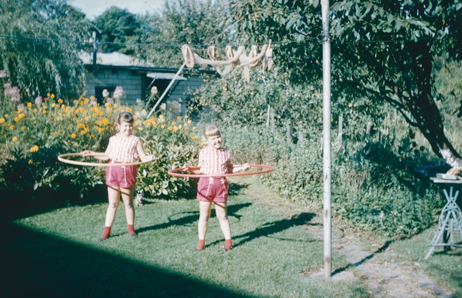 Two children are playing with hula hoops in a lush garden. They are wearing matching red and white striped shirts and red shorts. The garden is filled with colorful flowers and greenery, and there is a clothesline with items hanging in the background.