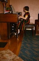 A person is seated at a wooden piano, attentively playing music with sheet music open in front of them. The individual is wearing a colorful striped dress and black shoes with white socks. The setting appears to be a cozy room with a blend of wooden and carpeted flooring. A variety of potted plants and decorative items sit atop the piano.