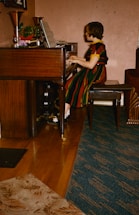 A person is seated at a wooden piano, attentively playing music with sheet music open in front of them. The individual is wearing a colorful striped dress and black shoes with white socks. The setting appears to be a cozy room with a blend of wooden and carpeted flooring. A variety of potted plants and decorative items sit atop the piano.